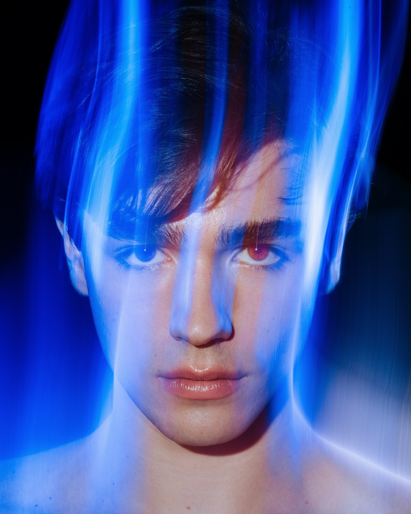 Portrait of a young adult male with dark hair, pale skin, and red eyes, illuminated with blue lighting and a black background.