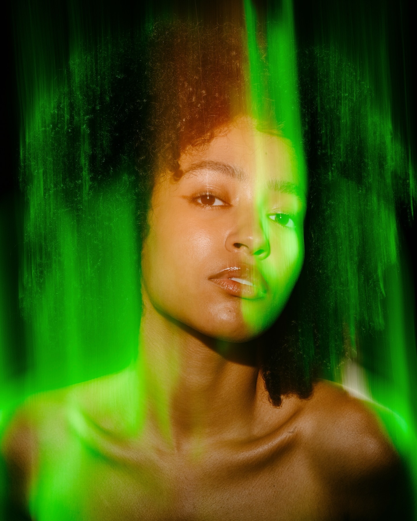 Portrait of a young woman with dark curly hair and bare shoulders, featuring diffused green lighting and a dark, shadowed background.