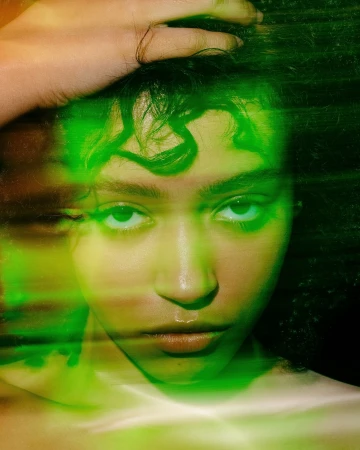 Portrait of a young woman with green lighting, hand on head, and soft focus. The girl is looking directly at the camera.