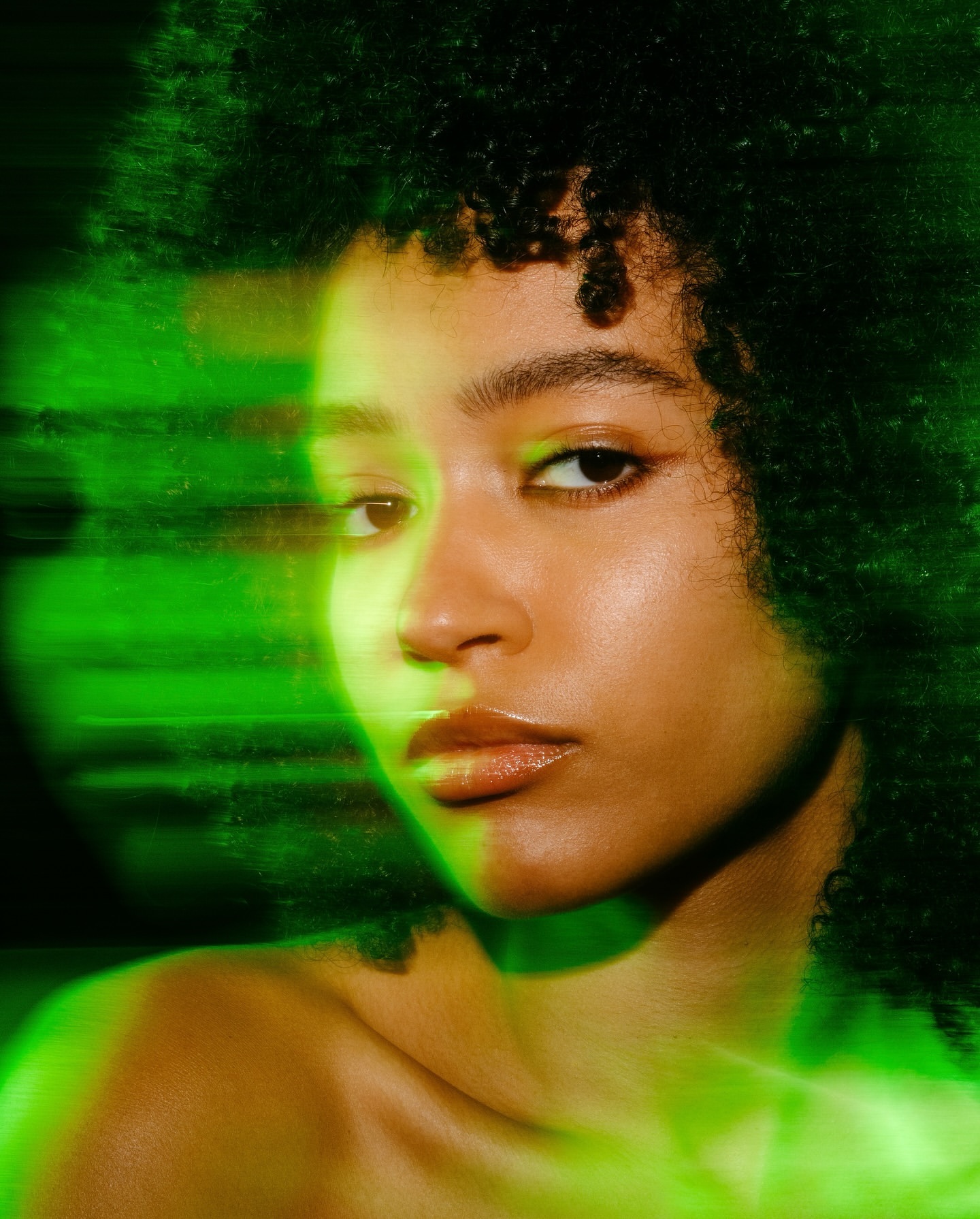 Close-up portrait of an adult woman with dark curly hair and tan skin, accented by green light on a dark background in a studio.