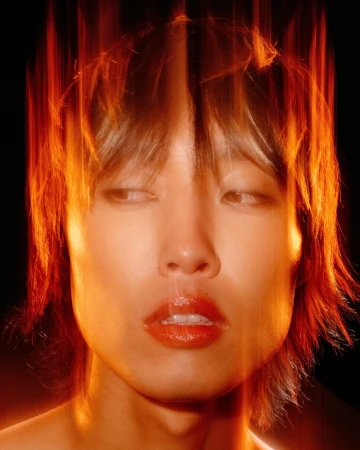 Close-up portrait of a person with glossy red lips and dark hair shot through vibrant orange light streaks by Derrick Freske