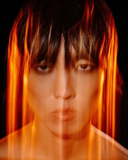 Close-up portrait of a woman with dark hair and light skin, highlighted by streaks of warm, diffused orange and yellow light.