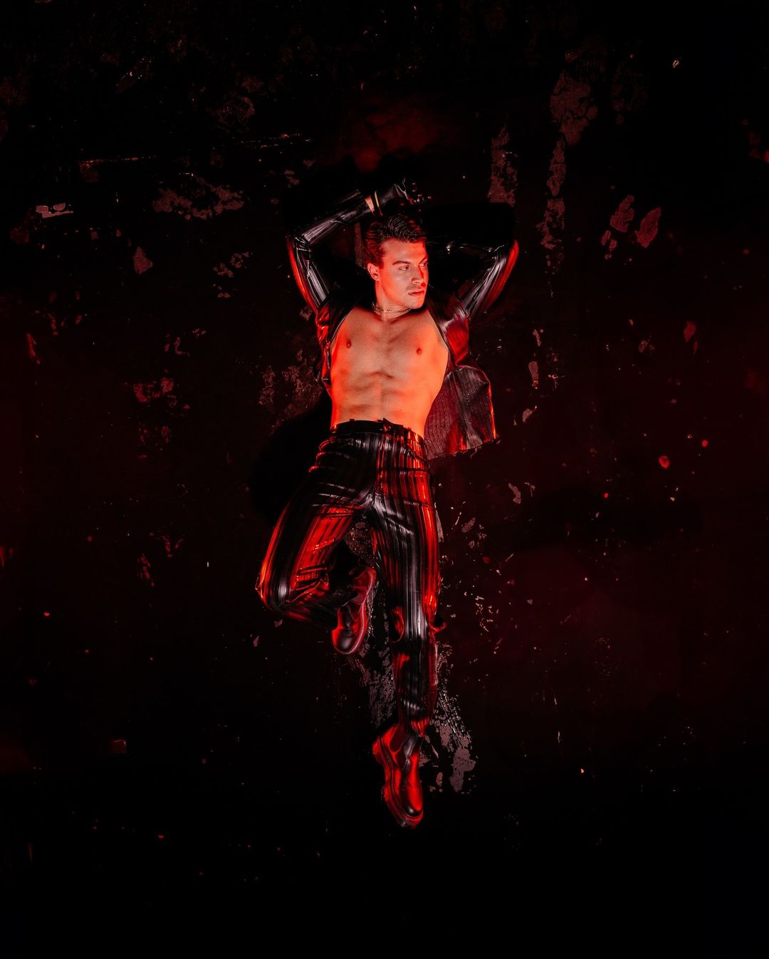 Male fashion model poses in shiny black vinyl top and pants on a mottled dark red background, lit from above for a studio shot.