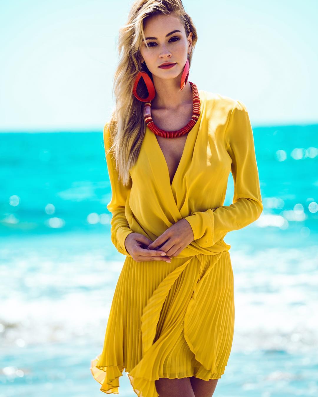 A blonde woman in a yellow dress stands on a beach with blue water in the background, wearing red earrings and necklace.