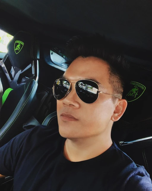 A man in aviator sunglasses sitting inside a luxury car with the Lamborghini logo visible on the headrest of the black leather seat.