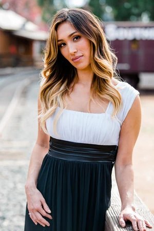 A woman with blonde-highlighted hair poses outdoors in a white and black dress, with railroad tracks visible in the soft-focus background.