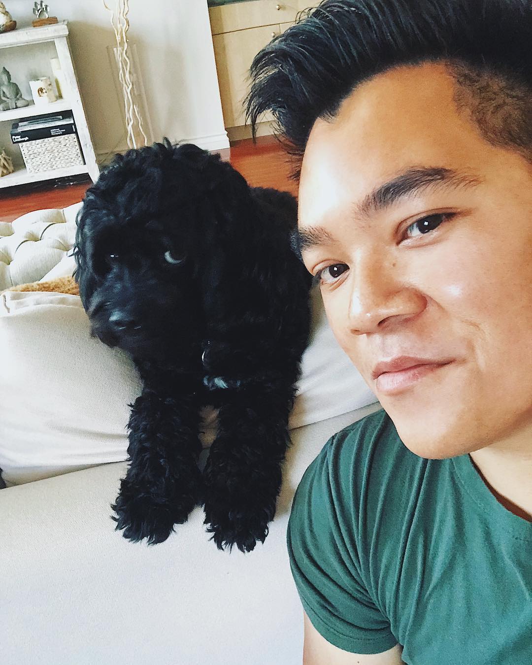 Close-up of a man and a black dog indoors. The man is wearing a green t-shirt, and the dog is lying on a white sofa.