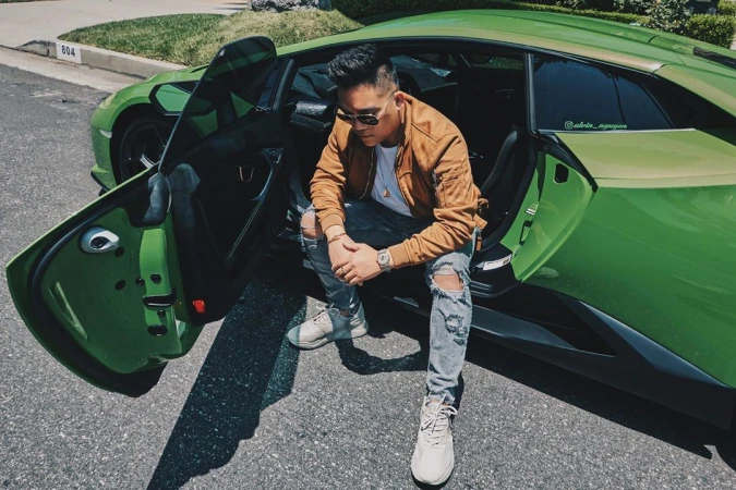 Adult male sits inside a green Lamborghini, wearing a jacket, ripped jeans and sneakers, posing on a paved street with houses in background.