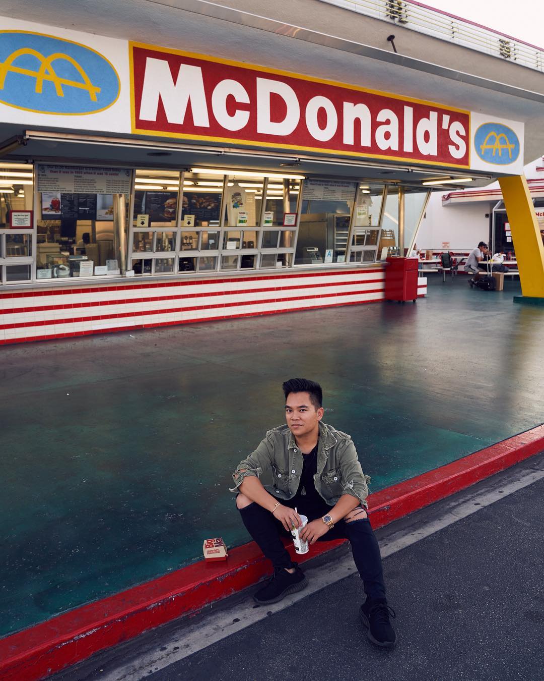 Man poses in front of a vintage McDonald's restaurant. He's seated on a ledge wearing ripped black jeans and an olive jacket.