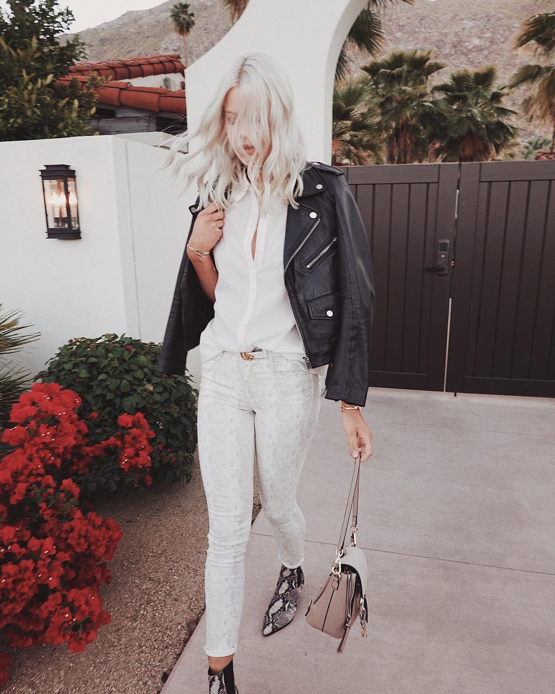 Blonde woman in black leather jacket and snakeskin boots walking outside a modern white building with palm trees and red flowering bushes.