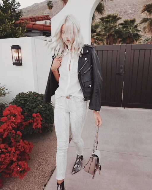 Blonde woman in black leather jacket and snakeskin boots walking outside a modern white building with palm trees and red flowering bushes.