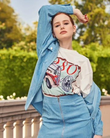A red-haired young woman poses outdoors wearing a blue corduroy skirt and jacket with graphic t-shirt, set against green foliage.