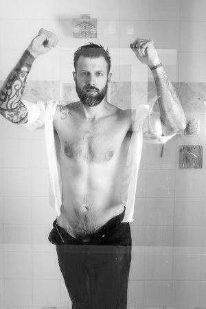 Monochrome portrait of a bearded man in a shower behind textured glass, wearing a shirt, arms raised, tattoos visible, tiled background.