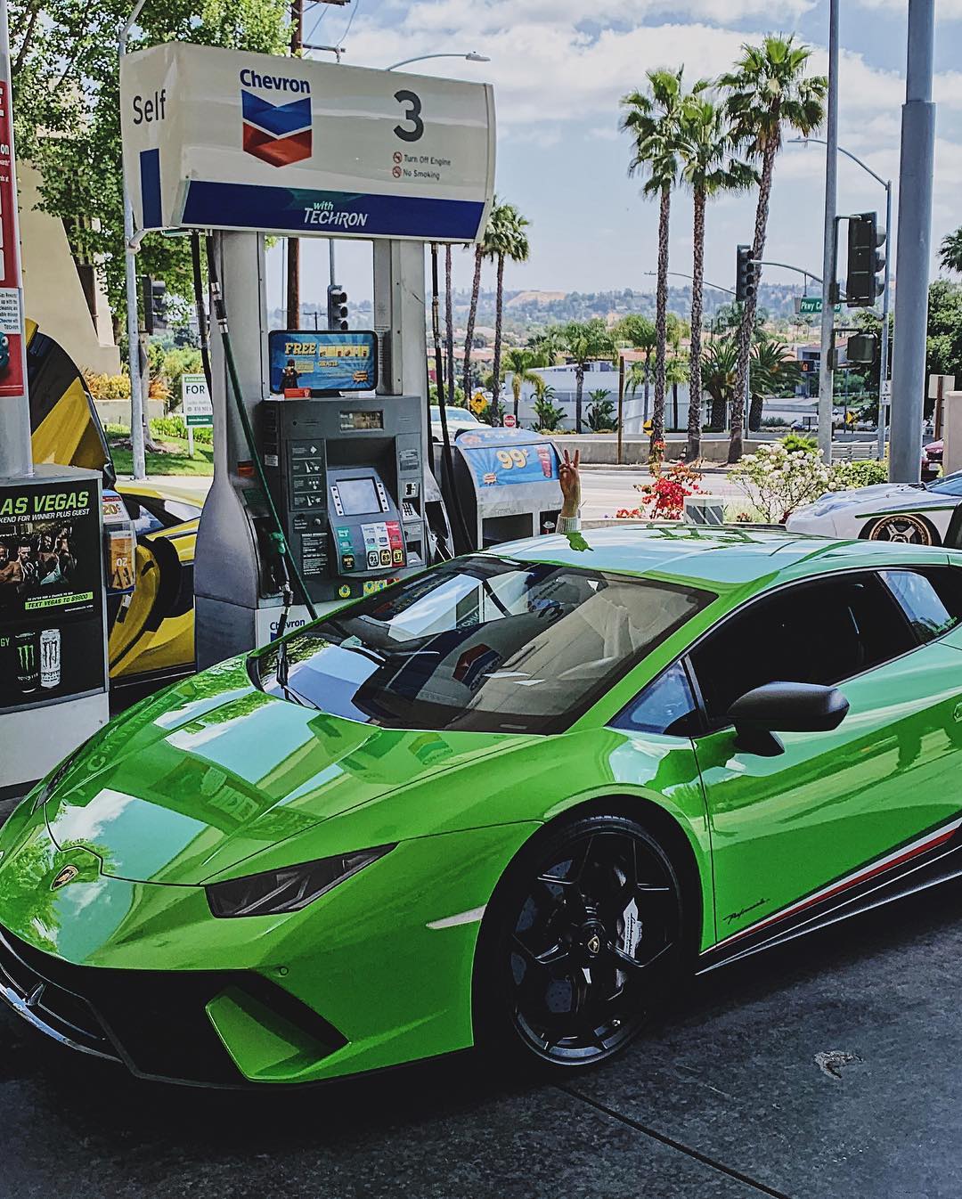 Green Lamborghini parked at a gas station, with palm trees and a hillside in the background, showcasing automotive design and sunny California lifestyle.