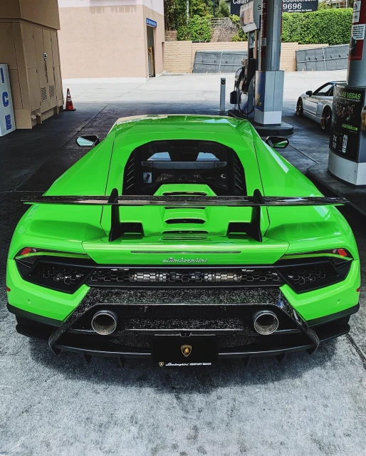 Rear view of a bright green Lamborghini Huracan parked at a gas station with fuel pumps and a glimpse of another car in the background.
