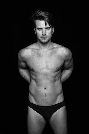 Striking black and white portrait of a shirtless adult male wearing briefs, posing against a stark black backdrop in a studio setting.