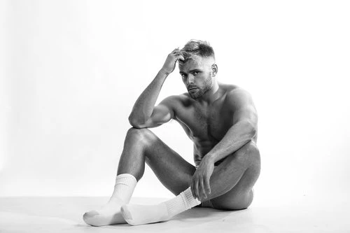Black-and-white studio shot of a shirtless adult male sitting on the floor wearing socks and looking at the camera with a neutral expression.