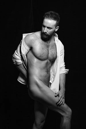 Black and white portrait of a bearded, muscular man wearing an open white shirt, posing shirtless against a dark background in a photo studio.