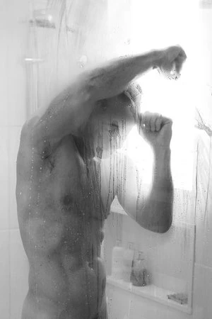 Monochrome image of an adult male in a steamy shower, arm raised, water droplets on glass, minimalist background, soft lighting, black and white.