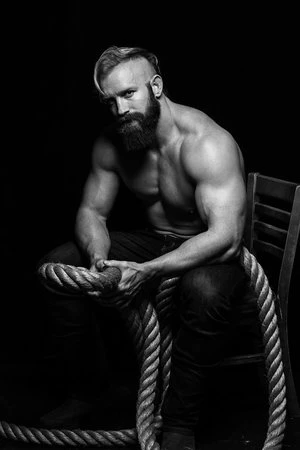 Striking black and white portrait of a muscular, bearded man with a mohawk, seated with a thick rope, against a dark backdrop.