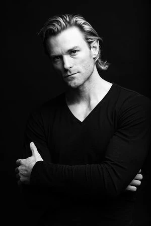 Portrait of a man with arms crossed wearing a black v neck shirt against a dark background