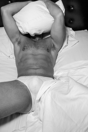Black and white studio shot of a muscular adult male lying in bed with white linens, holding a pillow over his head.
