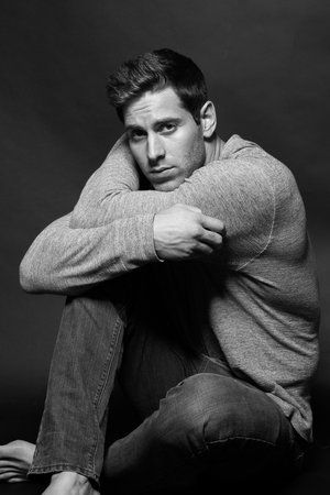 Monochrome studio shot of a male model sitting on the floor in a long-sleeve shirt and jeans, with his arms resting on his knees.