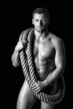 Shirtless man holding a thick coiled rope over his shoulder against a black background in a portrait by Alvin Nguyen