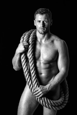 Shirtless man holding a thick coiled rope over his shoulder against a black background in a portrait by Alvin Nguyen