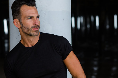 Portrait of an adult man with stubble wearing a black t-shirt, standing next to a white column in an indoor location.