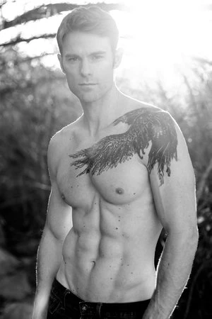 Monochrome portrait of shirtless man with abs and tattoo against blurred backdrop of foliage and bright sunlight, creating stark contrast.