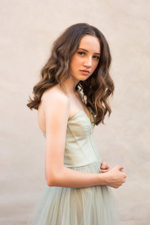 Portrait of a young woman with long curly brown hair wearing a light green strapless dress, posing against a neutral backdrop.