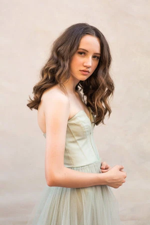 Portrait of a young woman with long curly brown hair wearing a light green strapless dress, posing against a neutral backdrop.