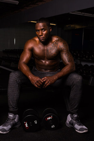 Muscular, shirtless adult man with tattoos sits on a bench with dumbbells in a gym with dark lighting, wearing joggers and athletic shoes.