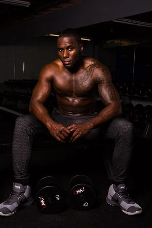 Muscular, shirtless adult man with tattoos sits on a bench with dumbbells in a gym with dark lighting, wearing joggers and athletic shoes.