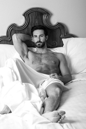 Black and white portrait of a shirtless man with a beard lying in a bed covered with a white sheet and pillows.
