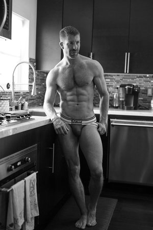 Black and white photo of muscular man in kitchen. Adult male with toned physique poses in underwear near counter and sink.
