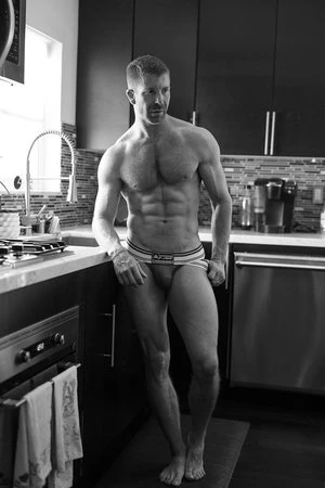 Black and white photo of muscular man in kitchen. Adult male with toned physique poses in underwear near counter and sink.