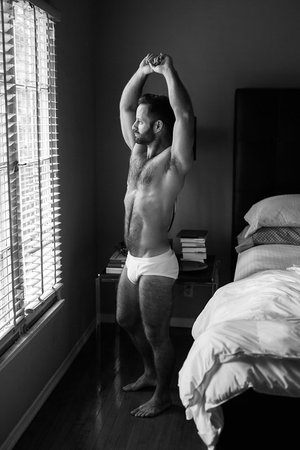 Black and white photo of a muscular man stretching arms upward toward a window in a bedroom with bed and bedside table.