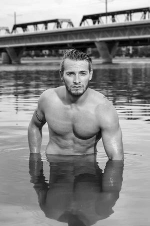 Black and white portrait of muscular adult male waist-deep in water with a beard and a bridge in the background on an overcast day.
