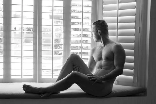 Monochromatic portrait of a shirtless adult male sitting on a window seat looking out through the shutters, high contrast lighting.