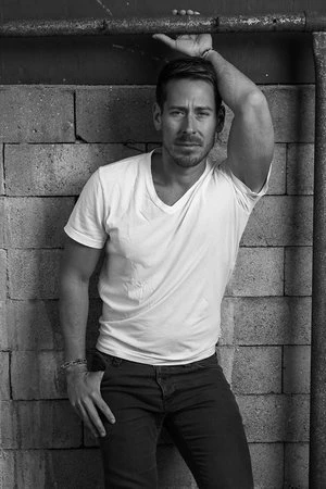 Monochrome portrait of a man in a white t-shirt leaning against a textured block wall with a metal pipe, looking at the camera.
