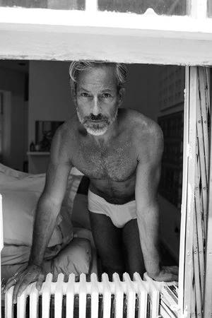 Monochrome portrait of shirtless gray-haired adult male in underwear peering from a window next to a radiator in an indoor setting.
