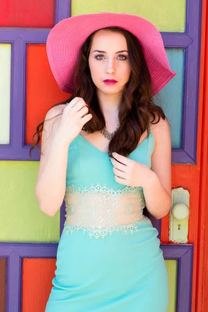 Portrait of a brunette woman in a turquoise dress and pink sunhat, posing against a multi-colored panel door in an outdoor setting.