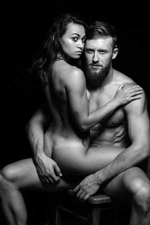 Black and white studio portrait of a nude mixed-race woman sitting on a stool, embraced by a bearded, nude man. Dark background.