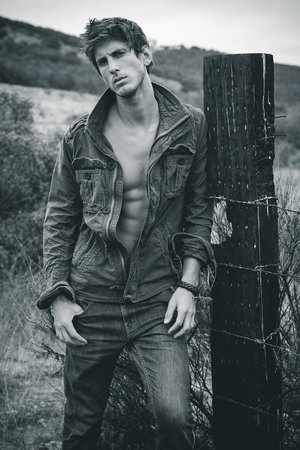 Monochrome portrait of a man with an unbuttoned jacket, leaning on a wooden post with barbed wire in an outdoor setting with a hillside backdrop.