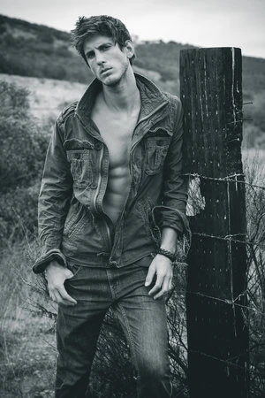 Monochrome portrait of a man with an unbuttoned jacket, leaning on a wooden post with barbed wire in an outdoor setting with a hillside backdrop.