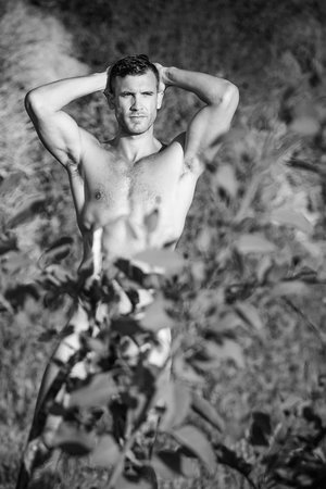 Black and white portrait of a muscular man posing outdoors with his hands behind his head, surrounded by leaves and greenery.