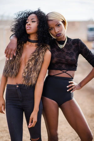 Two African-American women strike a fashionable pose outdoors, wearing black clothing with one in a fur vest, set against a desert-like backdrop.