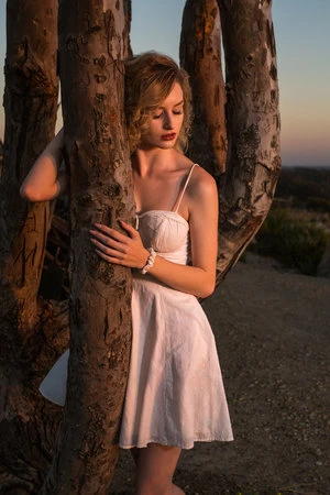 Blonde woman in a white dress leaning against a large tree trunk at sunset